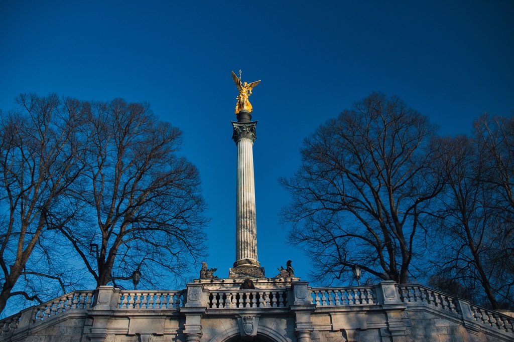 Der Friedensengel in München, eine goldene Engelsstatue auf einer hohen korinthischen Säule. Die Figur symbolisiert den Frieden und steht über einer steinernen Balustrade mit Treppen und Skulpturen. Links und rechts rahmen kahle Bäume die Szene ein, der Himmel ist klar und tiefblau.