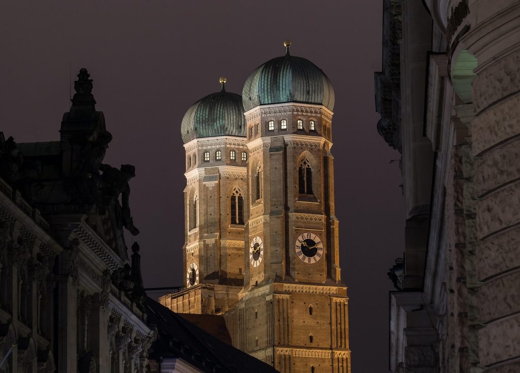 Die Frauenkirche in München bei Nacht, mit ihren zwei markanten Zwiebeltürmen aus Backstein, stimmungsvoll beleuchtet vor dunklem Himmel. Im Vordergrund sind die Fassaden umliegender Altbauten sichtbar.