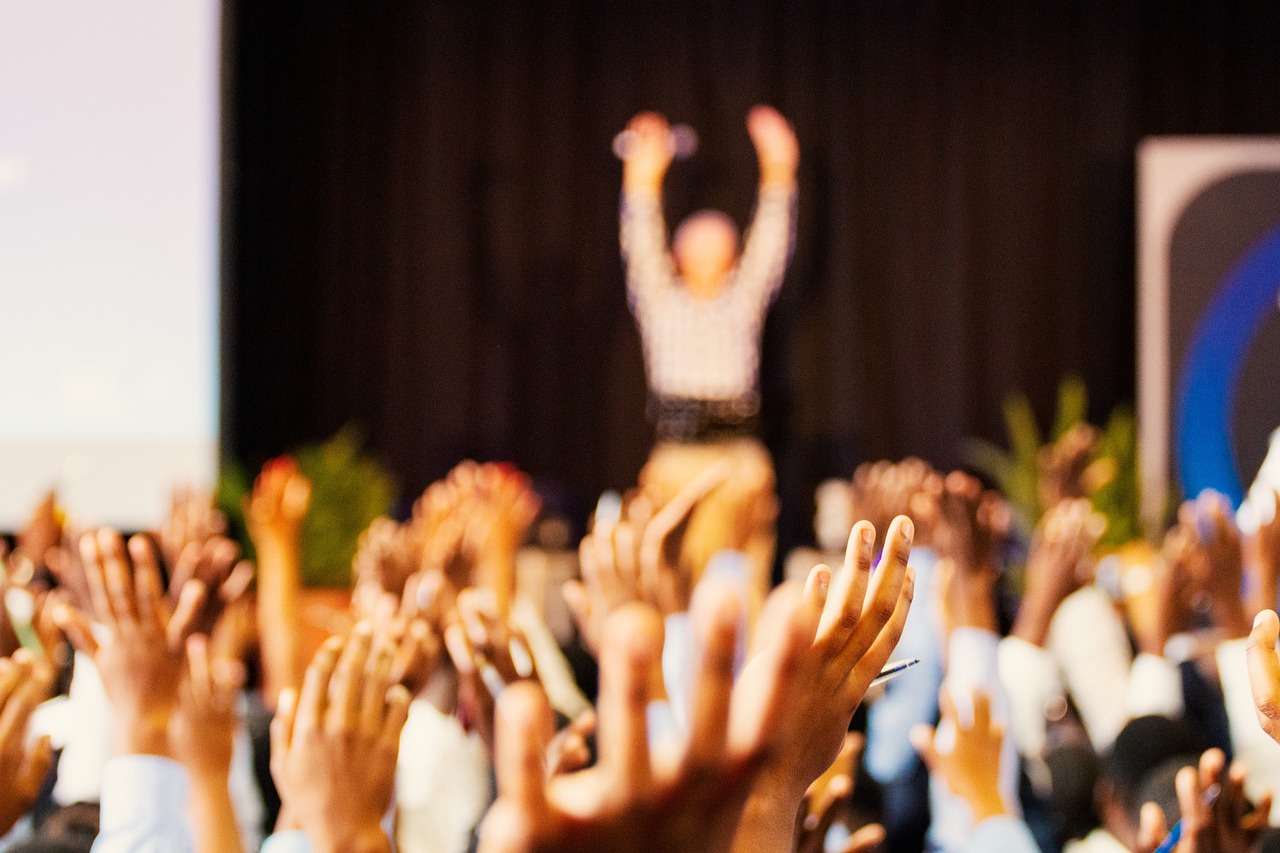 Großes Publikum mit erhobenen Händen vor einer Bühne, auf der eine Person begeistert spricht – symbolisiert die aktive Teilnahme der komenci GmbH im Bereich Performance Marketing.